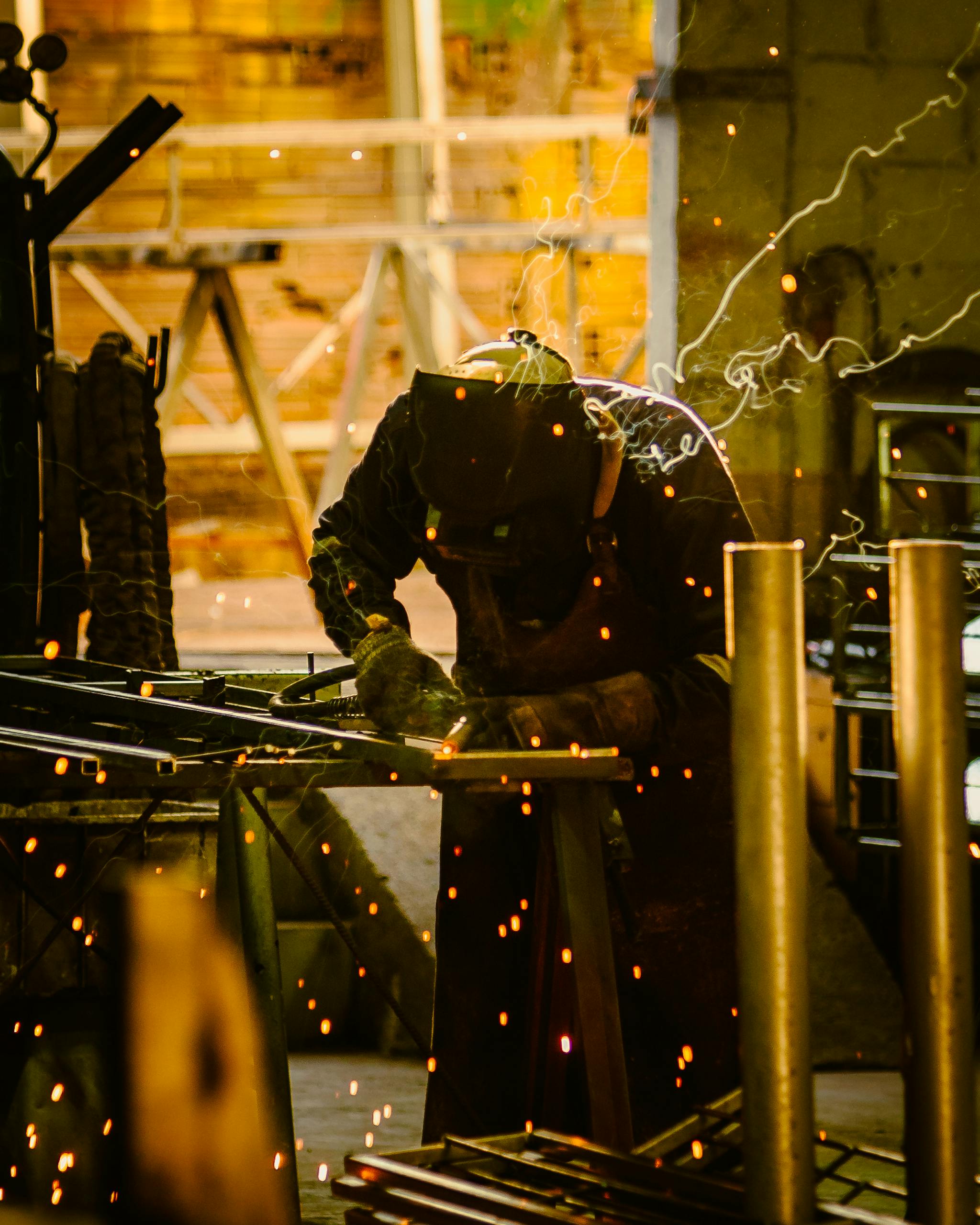 A welder using protective gear works on metal in a workshop, surrounded by bright sparks.